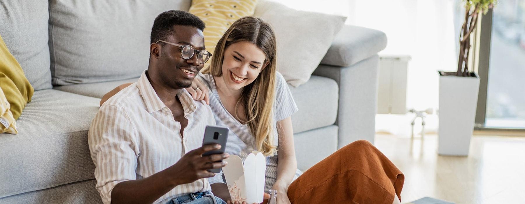 a man and woman with take out, sit against a couch on their living room floor and watch at their cell phone